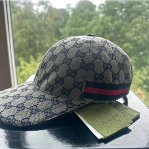 Gucci keps med GG-mönster och röd detalj - Pris kan diskuteras/ byten är intressantaSnygg keps från Gucci i beige och marinblått med klassiskt GG-monogram över hela kepsen. Kepsen har en böjd skärm och en ikonisk röd och marinblå rand på sidan. Tillverkad i bomullscanvas med justerbar rem baktill. Perfekt för dig som vill sticka ut med lyxig streetstyle.