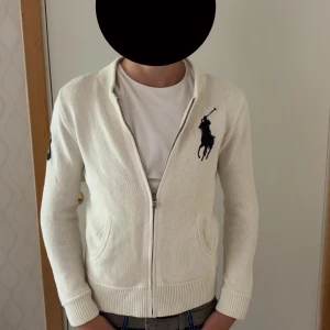 Vit stickad kofta från Ralph Lauren - Vit stickad kofta från Polo Ralph Lauren med dragkedja, ribbade muddar och två fickor fram. Stor broderad marinblå logga på bröstet och siffra 3 på ärmen. Perfekt för en clean och klassisk stil.