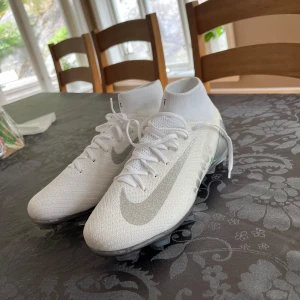 Vita Nike fotbollsskor med silvriga detaljer - Snygga vita Nike fotbollsskor med silvriga detaljer och glittrig swoosh. Skorna har högt skaft, snörning och en cool sula i krom och kolfiber-look. Ovandelen är i stickad syntet för en tight passform och maxad känsla på planen.