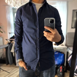 Mörkblå skjorta från Scotch & Soda - Snygg mörkblå skjorta från Scotch & Soda i klassisk modell med lång ärm och knappar hela vägen. Skjortan är tillverkad i bomull och har en stilren look som passar perfekt till jeans eller chinos. Märkesdetalj nertill och diskret stretch för extra komfort.