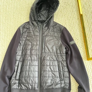 Svart quiltad jacka från Barbour - Snygg svart quiltad jacka från Barbour med huva och dragkedja. Jackan har släta ärmar, vadderad framsida och en diskret logga på ärmen. Perfekt för dig som gillar stilren och sportig look.