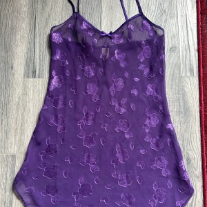 Lila blommig meshklänning med rosett - Superfin lila kortklänning i mesh med broderade blommor och smala axelband. Söt liten rosett framtill och transparent material som ger en trendig vibe. Perfekt för dig som vill sticka ut med en unik och lekfull look.