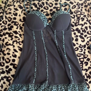 Svart bh/kortklänning med leoparddetaljer - Supersnygg svart bh/kortklänning med vadderade kupor och justerbara axelband. Klänningen har turkosa leopardmönstrade detaljer längs kupor, axelband, sömmar och volangkant nertill. Materialet är tunt och stretchigt, perfekt för att sticka ut.