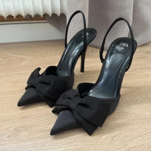 Svarta pumps med rosett från Zara - Stilrena svarta pumps från Zara med spetsig tå och hög klack. Skorna har en elegant rosett framtill och smal rem runt hälen. Tillverkade i ett matt tyg. Nyskick. Storlek 38.