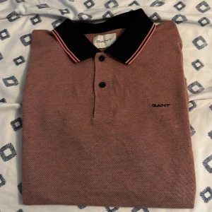 Rosa pikétröja från GANT med svart krage - Säljer en snygg rosa pikétröja med mörkblå detaljer imellan som sitter perfekt på den här piken från GANT med mörkblå krage och rosa ränder. Tröjan har två knappar framtill och diskret GANT-logga broderad på bröstet. Perfekt för en stilren och avslappnad look. Som är köpt från Gant butiken i NK för 1000 piken är knappt använd och säljs för 500 i storlek M men passar L också 