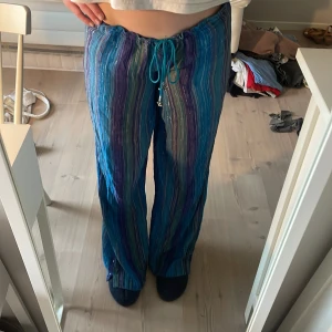 Lågmidjade blå-lila linnebyxor med snörning - Säljer ett par färgglada lågmidjade linnebyxor med blå, lila och gröna ränder. Byxorna har relaxed fit, raka ben och dragsko i midjan för skön passform. Perfekta för varma dagar när du vill ha något luftigt och snyggt. De är i storlek L men går att spänna åt mycket i midjan.