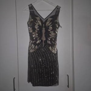 Paljettklänning gatsby - En svart klänning med glittrande detaljer i guld. Klänningen har en V-ringning och är figursydd. Den är perfekt för en festlig tillställning.  Snygg klänning med glittrande detaljer och ett vackert fjärilsmönster. Klänningen har en v-ringning och är figursydd.