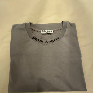 Grå Palm Angels t-shirt med tryck - Helt ny och oanvänd grå t-shirt från Palm Angels med svart logotyptryck både fram vid halsen och stort på ryggen. Klassisk rund halsringning och korta ärmar. Skön och mjuk bomullskvalitet, perfekt för en clean streetwear-look.