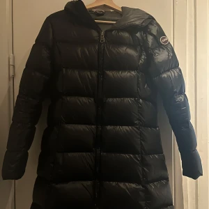  Colmar dunjacka  - Säljer en svart, lång och quiltad dunjacka från Colmar. Jackan har huva, dragkedja framtill och diskret logga på ärmen. Perfekt för kalla dagar med sin puffiga look och stilrena design. Storlek 38 