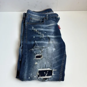 Dsquared2 blå slitna jeans med färgstänk - Riktigt snygga blå jeans från Dsquared2 med coola slitningar, hål och vita färgstänk. Jeansen har en smal passform, klassisk femficksdesign och tydliga tvättade detaljer. Perfekta för dig som gillar en edgy streetstil.