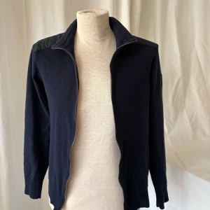 Mörkblå cardigan från Belstaff - Snygg mörkblå cardigan jacka från Belstaff med quiltade detaljer på axlarna och dragkedja framtill. Jackan är i mjuk ullmix och har en klassisk passform med ribbade muddar. Strl M men sitter som S.
