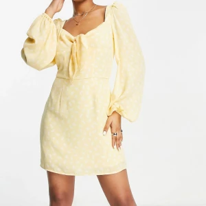 Gul blommig kortklänning  - Superfin ljusgul kortklänning med vita små blommor, långa puffärmar och knytning framtill. Klänningen har fyrkantig urringning och är figursydd i midjan. 