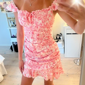 Rosa blommig offshoulder kortklänning - Supersöt kort klänning med rosa blommigt mönster och offshoulder-ärmar. Klänningen har volangkant nertill, smockad passform och knytning framtill. Perfekt för sommaren