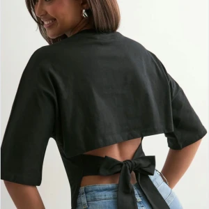 Svart croppad topp med knyt i ryggen - Svart  topp med halvlånga ärmar. Baksidan har en öppen design med breda band som knyts i en stor rosett i ryggen. nelly nyskick 