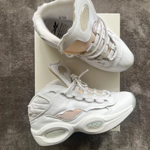 Maison Margiela x Reebok Question Mid - Unika vita sneakers från Maison Margiela och Reebok med högt skaft, chunky sula och genomskinliga detaljer. Skorna har snörning, inslag av beige och coola paneler i skinn och syntet. Perfekta för dig som vill sticka ut med en futuristisk vibe.