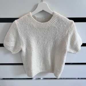 Vit stickad tröja från Gina Tricot - Vit stickad tröja från Gina Tricot med korta ärmar och ribbad rund hals. Tröjan är lite croppad och har en fluffig känsla, perfekt att styla med jeans eller kjol för en chill look.
