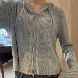 Grå ribbad hoodie med dragkedja Monki - Grå ribbstickad hoodie från Monki i storlek M, passar även XS och S. Tröjan har dragkedja framtill, huva med snörning och långa ärmar 💕Materialet är mjukt och stretchigt med 76% viskos och 24% polyester. Perfekt till chill dagar och enkel att matcha med jeans! :)