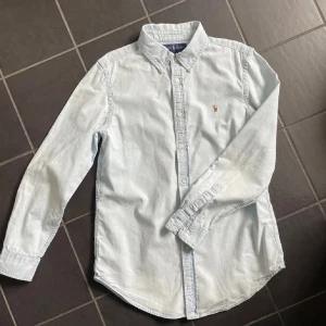Ljusblå skjorta från Ralph Lauren - Storlek L i barn så skulle säga S. Klassisk ljusblå skjorta från Ralph Lauren med button down-krage och broderad logga på bröstet. Skjortan är långärmad, har normal passform och är tillverkad i mjuk bomull. Perfekt för en clean och stilren look.