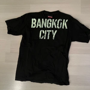 Svart t-shirt med Bangkok City-tryck - Svart vintage t-shirt från United CGBG med stort vitt 'BANGKOK CITY'-tryck på bröstet och mindre tryck på ryggen. Storlek L/XL, pris 150 kr 