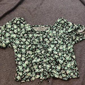 Blommig blus med snörning - Supersöt blommig blus i svart med gröna och vita blommor. Toppen har korta puffärmar, smockad rygg och snörning framtill för extra detalj. Perfekt för dig som gillar romantisk och trendig stil.
