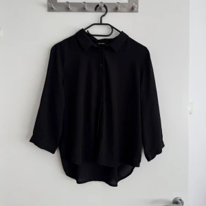 Snygg svart skjorta från Monki - Stilren svart skjorta från Monki i 100% polyester. Skjortan har klassisk krage, knappar framtill och trekvartsärmar. Modellen är lite längre baktill för en snygg och avslappnad look. Perfekt att styla med jeans eller kjol.