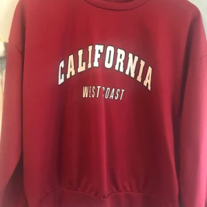 Röd sweatshirt med California-tryck - Cool röd sweatshirt med trycket 'CALIFORNIA WEST COAST' i vitt och svart på bröstet. Tröjan har rund halsringning och långa ärmar, perfekt för en avslappnad och trendig look. Mjuk och skön, passar till jeans eller leggings.
