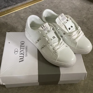 Valentino Rockstud vita sneakers - Snygga vita Valentino Rockstud sneakers med ikoniska silverfärgade nitar och klassisk snörning. Skorna är i skinn och har en platt sula med rund tå. Perfekta för dig som vill ha en clean men edgy look.