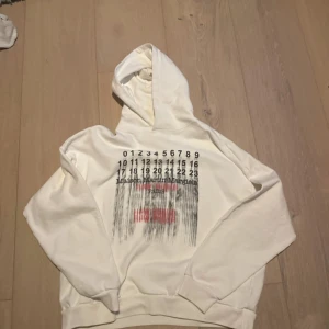 Vit hoodie Maison Margiela print ALDRIG ANVÄND - Vit hoodie från Maison Margiela med ikoniskt siffertryck och svart/röd grafisk print på bröstet. Klassisk huva, ribbade muddar och relaxed fit. Perfekt för dig som gillar streetstyle och vill sticka ut med en unik designertröja.