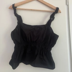 Svart linnetopp med twistade axelband - Säljer en svart linnetopp med twistade axelband och smockad midja. Toppen har en luftig passform och är perfekt för varma dagar. Materialet känns lätt och toppen har en enkel men trendig look.