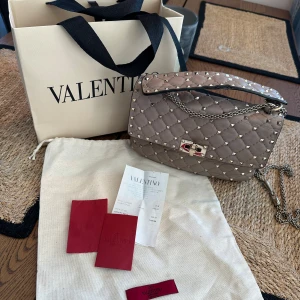 Valentino Garavani väska - Valentino Garavani Rockstud spike leather bag i färgen poudre.   Säljer min ikoniska Valentino Garavani Rockstud Spike-väska i en ljusbeige/ljus rosa färg .Väskan är tillverkad i quiltat kalvskinn och prydd med de klassiska pyramidformade nitarna. Den har en elegant kedjerem som kan bäras både dubbelt på axeln eller enkel som crossbody. Detta band går även att ta bort.   Väskan är använd i 4 år där lädret har fått visst slitage. Se alla bilder. I övrigt är det ingen fel på väskan och låset är pr
