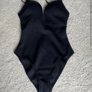 Helt ny Svart baddräkt från Hunkemöller S - Supersnygg svart baddräkt från Hunkemöller med vadderade kupor och smala axelband. Snyggt formad med en liten v-ringning framtill och guldfärgat spänne i ryggen. Perfekt för stranden eller poolen i sommar!