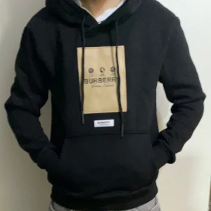 Svart Burberry hoodie med beige patch - Svart hoodie från Burberry med stor beige patch och logga på bröstet. Med en luva och snöre. Mjuk bomullskänsla och relaxed fit, perfekt för en chill och stilren look.
