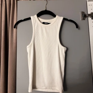 Vit ribbad linnetopp från Bik Bok - Säljer en stilren vit ribbad linnetopp från Bik Bok i storlek S. Toppen är ärmlös med klassisk rund halsringning och har en tight passform. Perfekt att styla med jeans eller kjol för en clean look.