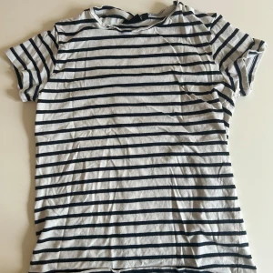 Randig t-shirt från KappAhl XS - Snygg randig t-shirt från KappAhl i storlek XS. T-shirten är vit med mörkblå horisontella ränder och har en klassisk rund halsringning samt korta ärmar. Perfekt för en chill och stilren look. Materialet är mjuk bomull som känns skönt mot huden.