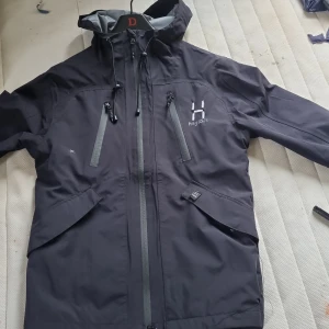 Haglöfs jacka vind - Snygg svart vindjacka från Haglöfs med grå detaljer och justerbar huva. Jackan har flera dragkedjeförsedda fickor, reflexdetaljer på ryggen och är perfekt för blåsiga dagar. Lätt och smidig design med hög krage och smarta funktioner.