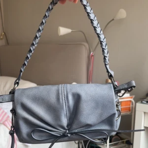 Grå axelväska med flätat handtag - Snygg grå axelväska i skinnimitation med flätat handtag och dekorativ knytdetalj framtill. Väskan har en mjuk, rundad form och metallfästen på sidorna. Perfekt för att lyfta din outfit med en cool touch.