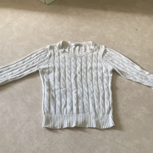 Vit kabelstickad tröja Gina Tricot XS - Vit kabelstickad tröja från Gina Tricot i storlek XS. Tröjan har ett klassiskt kabelmönster, rund halsringning och ribbade muddar vid ärmslut och nederkant. Perfekt för lager-på-lager och enkel att matcha med jeans eller kjol 