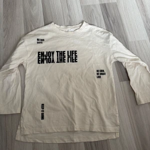 Beige långärmad tröja med tryck  - Beige långärmad tröja med coola svarta tryck som 'ENJOY THE LIFE' och 'NO BAD DAYS'. Tillverkad i mjuk ekologisk bomull. Tröjan har slits i sidorna och en avslappnad passform, perfekt för en chill och stilren look.