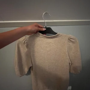 Beige puffärm tröja från 157 - Superfin beige tröja från 157 med puffärmar och rund hals. Tröjan har en mjuk känsla och är perfekt om du vill ha en enkel men ändå trendig look. Passar till både jeans och kjol och är lätt att matcha med andra plagg.