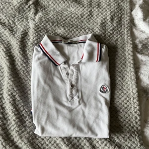 Vit pikétröja från Moncler S - Vit pikétröja från Moncler i bomull med klassisk krage och knäppning framtill. Kragen har röda och marinblå ränder för en sportig vibe. Broderad Moncler-logga på bröstet och korta ärmar. Perfekt för dig som gillar stilrena och exklusiva plagg.