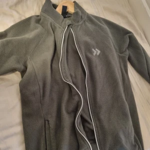 Grå fleecejacka från 365 Funk - Mysig grå fleecejacka från 365 Funk med hög krage och hel dragkedja. Jackan har två sidofickor och en enkel, stilren design med en liten logga på bröstet. Perfekt för kyliga dagar när du vill hålla dig varm och bekväm.