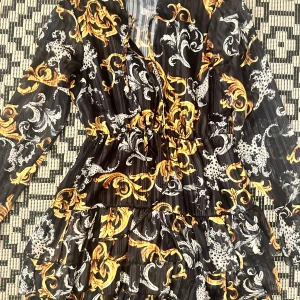 Svart och guldig mönstrad kortklänning - Snygg kortklänning med svart bas och detaljerat mönster i guld och vitt. Klänningen har långa ärmar, omlottliv och volang i kjolen. Materialet är lätt och genomskinligt, perfekt för dig som gillar statement-plagg med lyxig känsla. Går över knäna . Aldrig använd