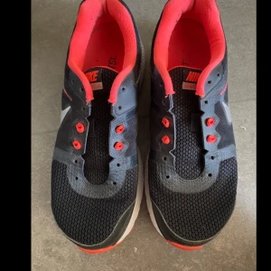 Svarta Nike sneakers med röda detaljer - Snygga svarta Nike sneakers med mesh-ovandel, röda detaljer och vit sula. Skorna har en sportig look med silverfärgad swoosh på sidan.