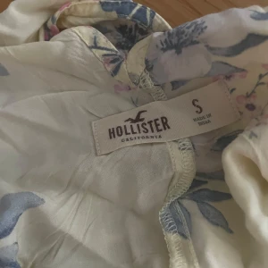 Blommig linnetopp från Hollister - Supersöt ljusgul linnetopp från Hollister med blommigt mönster i blått och rosa. Toppen har tunna axelband och en luftig passform, perfekt för varma dagar. Materialet känns lätt och somrigt, och den blommiga looken ger en fräsch vibe.