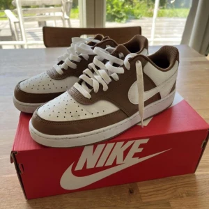 Nike Court Vision sneakers brun/vit - Nike Court Vision sneakers i snygg brun och vit färg. Klassisk låg modell med perforerad tå, vit sula och bruna detaljer. Skorna har snörning och är tillverkade i syntet och skinn. Perfekta för dig som gillar retrovibbar och stilren design. Helt nya Nike sneakers säljes. Använda endast en gång men var för små. unisex stl 39. Inköpta på stadium i juni. 