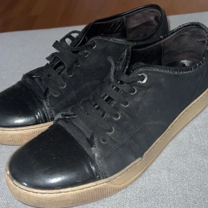 Svarta lanvins - Snygga svarta sneakers från Lanvin med klassisk design. Skorna har svart mocka på ovandelen, lackad svart tå och svarta skosnören. Sulan är beige och ger en schysst kontrast. Perfekta för dig som gillar stilrena och exklusiva sneakers. Stormed 42 men Passar även 43 och 44 