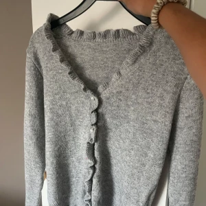 Grå kofta med volang och pärlknappar - Superfin grå kofta med volangkant och pärlknappar. Mjuk och stickad. Långärmad och passar till både jeans och kjol. Nyskick