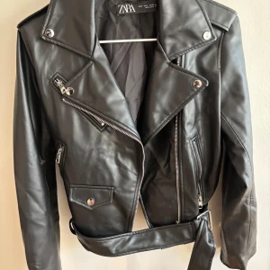 Svart skinnjacka från Zara, M - Snygg svart skinnjacka från Zara med klassisk bikerstil, silvriga dragkedjor, breda slag och bälte i midjan. Jackan har flera fickor med dragkedja och tryckknappar som ger en cool vibe. Perfekt för dig som vill ha en edgy look.