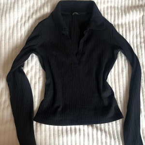 Svart ribbad pikétröja från Monki - Svart långärmad pikétröja från Monki med ribbad struktur och  krage. Tröjan är tight och har djup v-ringning. Tröjan ser både professionell och elegant ut så passa på att se classy ut i sommar med denna💗