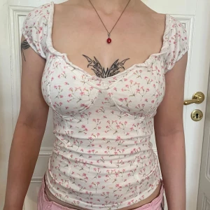 Söt blommig topp med volangkant - Supersöt vit topp med små rosa blommor, volangkant vid urringningen. Figurnära passform och mjukt bomullsmaterial. Perfekt för en romantisk och feminin look.